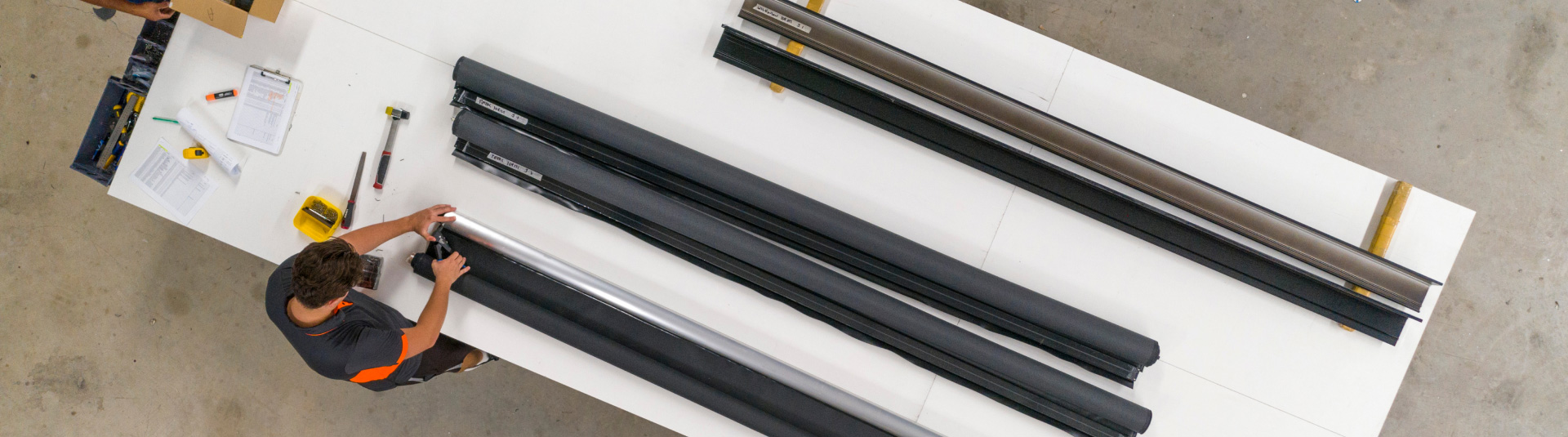 Person working on long cylindrical parts on a white table with tools and documents in a workshop.