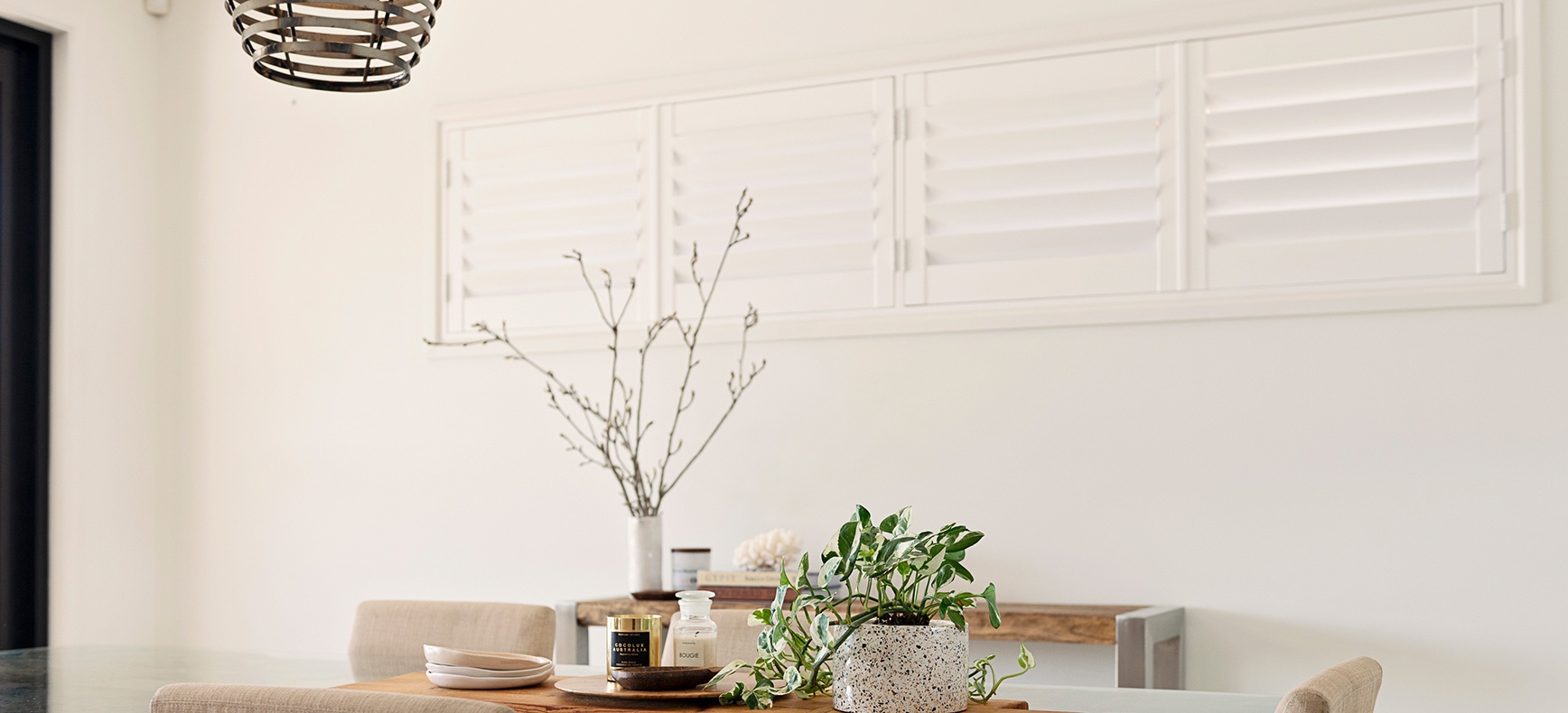Minimalist dining area with wooden table, beige chairs, plant pot, candles, and sideboard.